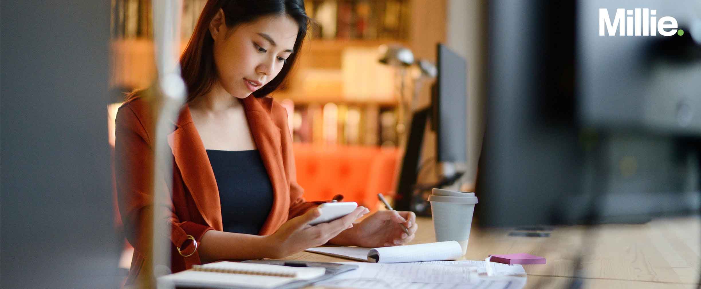 Photo of a young women sitting at a desk with a pen and pad of paper, reviewing her personal finances on her phone.