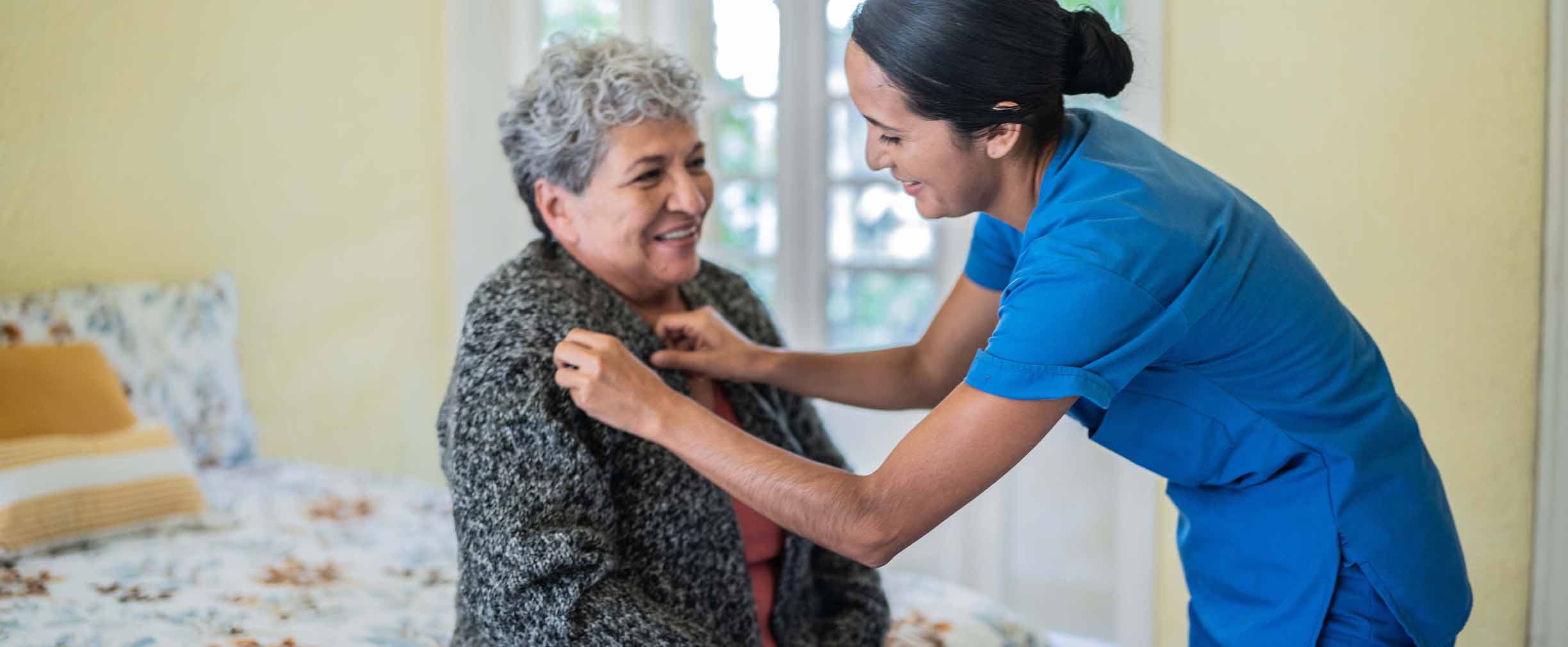 A health care worker in blue scrubs laughs with an older woman as she helps put on the woman’s sweater 