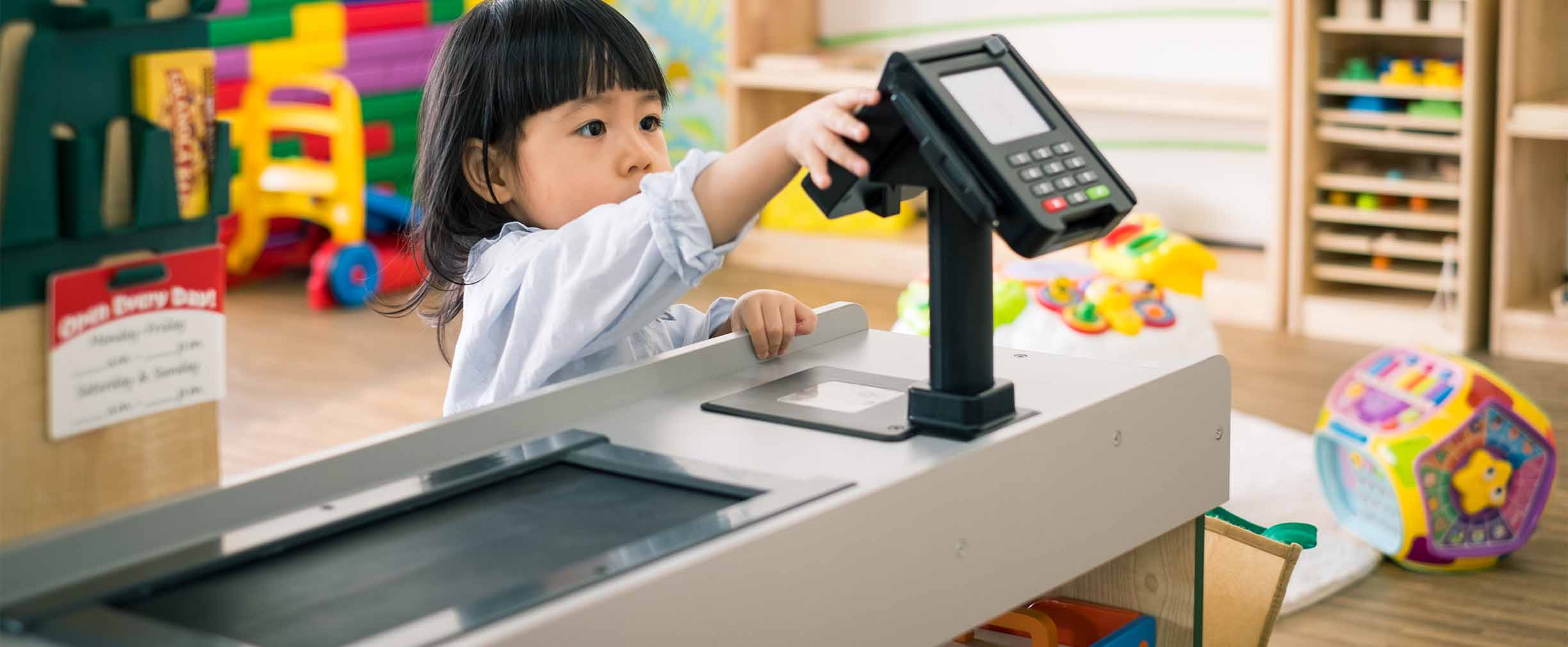 A kindergarten-aged girl plays with the credit card scanner on a play grocery store checkout counter 