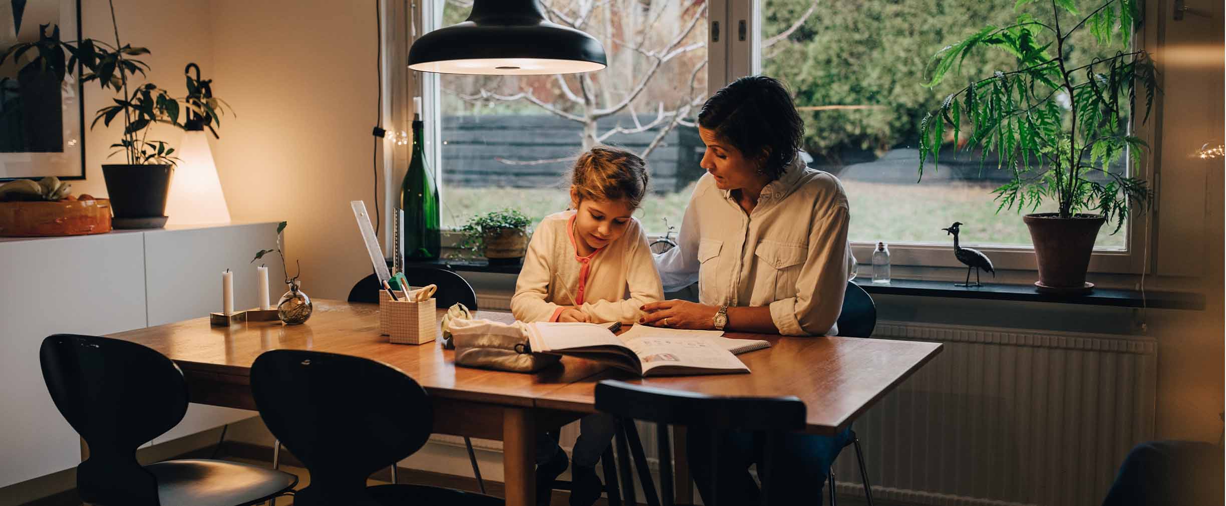 In a plant-filled dining room, a parent or grandparent helps a young girl work on her homework